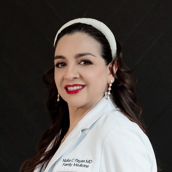A woman with long dark hair, wearing a white headband, pearl earrings, and a white lab coat with the text Nidia C. Payan MD Family Medicine, poses against a dark background and smiles at the camera.